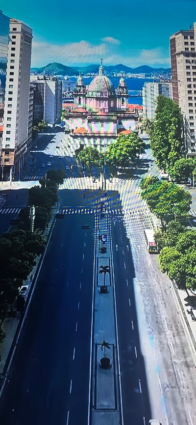 The image displays an aerial view of a wide, empty street in a city, likely a harbor city, given the presence of water and hills in the background. Dominating the center of the frame is a large, ornate building with a dome and several smaller cupolas, suggesting it is a significant religious or governmental structure. Flanking the street are tall buildings, some with modern glass facades and others appearing older and more ornate. Lush green trees line portions of the street, providing shade and breaking up the urban landscape. The street itself is mostly devoid of traffic, with only a few scattered vehicles visible, indicating a quiet or perhaps restricted period of activity. The sky is a clear blue with a few wispy clouds, and the sun casts long shadows across the pavement.

There is no indication that this image is of Lippstadt, Germany. The architecture and landscape are not consistent with Lippstadt's known geography and urban development. The presence of a large domed building reminiscent of European religious architecture, the hilly coastline, and the overall urban density do not align with Lippstadt's characteristics.