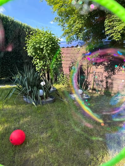 This image depicts a backyard scene in Lauchhammer, Germany. A section of green lawn occupies the lower-right area, upon which lies a red rubber ball. An ornamental garden bed with green plants and two white spherical garden lights is situated towards the left. A weathered wooden fence stands in the background, visible through a partially obscured section, potentially due to a camera lens artifact. A green bubble wand frame appears partially in the shot, along with a rainbow-colored reflection, possibly created by the wand’s soapy film. The lighting suggests a sunny day. The presence of the ball and garden elements indicate a residential backyard setting.