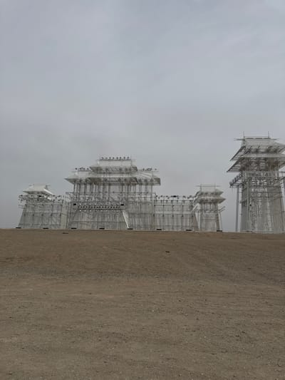 The image shows the Yadan World Geopark in Guazhou County, China. The park features unique geological formations resembling buildings or structures arranged along a flat, desert-like landscape under an overcast sky. The formations are primarily light-colored, with complex frameworks of what appears to be interconnected scaffolding or pipe-like structures. The foreground consists of dry, compacted earth.
