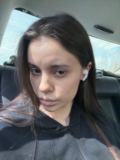 A close-up, tilted portrait captures a young woman inside a car, likely in Ustinovo, Russia, during daytime. She has long, dark brown hair with visible lighter highlights and is wearing a dark, possibly black, zippered top. A white wireless earbud is placed in her right ear. She looks directly towards the viewer with a neutral, slightly serious expression; her lips are slightly parted.
Strong natural light enters the car from behind and to the side, creating distinct shadows across her face and body. The background reveals a car interior with dark upholstery and the rear window, showing clear defroster lines and bright, possibly sunny, outdoor conditions. A small, dark, elongated object is affixed to the car's ceiling or interior trim above her head. No text is visible in the image.