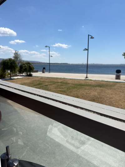 The image depicts a waterside scene, likely in Çanakkale Merkez, Turkey, viewed from an interior space. In the foreground, a metallic railing with a textured surface is visible, suggesting the perspective is from a cafe, restaurant, or balcony. Beyond the railing, a grassy area transitions to a paved walkway bordering a body of water.

Several lampposts with dual lights are spaced along the walkway. Two cylindrical trash receptacles are also present. In the middle ground, two rounded trees with lush foliage stand on the grassy area. The body of water, likely a sea or large bay, occupies the background, with a hazy shoreline visible in the distance under a bright blue sky dotted with scattered white clouds. The overall impression is a serene, daytime outdoor environment.