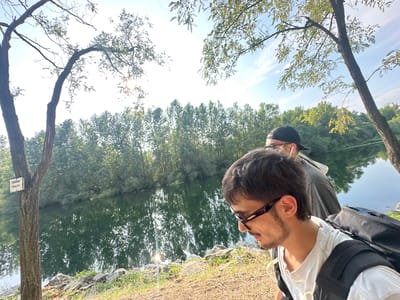Two people are standing by the side of a calm body of water, with trees and foliage lining the opposite bank. The person in the foreground, a young man with dark hair and a goatee, wears sunglasses and a white t-shirt. He has a backpack on, with a blue strap visible. Behind him, another person wearing a black baseball cap and glasses is partially visible. The scene appears to be outdoors, with sunlight filtering through the trees, suggesting it's daytime. The water reflects the trees and sky. A small sign on a tree trunk in the left foreground reads "TENERE PULITO," which translates to "KEEP CLEAN" in Italian, providing a location cue for Rudiano, Italy. The overall atmosphere seems relaxed and peaceful.