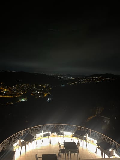 The image depicts a night scene at an elevated outdoor seating area overlooking a city at the foot of a mountain range. The foreground features a curved balcony with several tables and chairs arranged for outdoor dining. The tables are dark and square, and the chairs are simple, dark, metal frames with seats and backs. A decorative white railing curves along the edge of the balcony. The lighting on the balcony is a warm, yellowish hue, illuminating the floor and furniture. Beyond the balcony, the landscape descends into darkness, with a sprawling city illuminated by a multitude of lights. The city lights are concentrated in clusters and appear to follow the contours of the terrain, suggesting a hilly or mountainous urban environment. Distant hills and mountains are visible against the night sky, with faint lights indicating further settlements or structures. The sky is dark with some diffused light, possibly from clouds or distant light pollution. The overall impression is of a scenic viewpoint offering a panoramic vista of a city at night. No people are visible in the image. The scene appears to be a restaurant or cafe terrace.