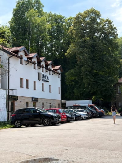 A vertical shot shows a white building with a dark, angular roofline, likely a hotel, with the text "JUNCA HOTEL" prominently displayed. To the right of the building, a line of parked cars, including a black SUV, a red car, and a white van, are visible. Further to the right, a person with long dark hair wearing a black top and denim shorts stands on a paved surface, casting a shadow. A large tree with lush green foliage dominates the left side of the frame, with a bright blue sky and scattered clouds above. The scene appears to be set during the daytime, under sunny weather conditions, possibly in Jajce, Bosnia and Herzegovina. The overall impression is a typical outdoor scene with a hotel and parking area surrounded by nature. The sign "RESTORAN" is also visible on the building.