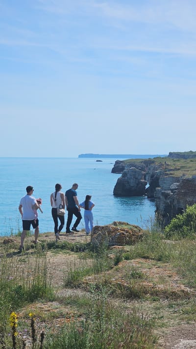 Four people stand on a cliff overlooking the Black Sea in Kamen Bryag, Bulgaria. A man in a white shirt and black shorts faces left, holding a child. Behind them, a woman in a white shirt and black pants faces left. Behind her, a man in a green shirt and black pants faces left. At the edge of the cliff, a woman in a blue shirt and light blue pants faces left, looking at the sea. Behind them are cliffs, the sea, and the sky. The vegetation on the cliff is dry grass. There are rocky formations in the sea.