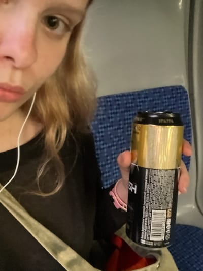A close-up, slightly blurry, medium shot shows a person holding a black and gold can of energy drink. The person, whose face is partially visible in the upper left, has fair skin, light brown hair, and is wearing a black top. A white earphone wire is visible. The can, held in the person's right hand, has Russian text and a barcode on its black portion. The background features a blue patterned seat, likely in a public transportation setting. The image does not contain any specific identifiable landmarks or features of Kokkorevo, Russia.
