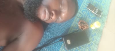 A dark-skinned man with a beard is lying on his side on a blue and green patterned mat. His eyes are closed. Near him are a black mobile phone, a broken mobile phone with a rubber band around it, a string of brown beads, and a small yellow container. The location is speculated to be Bende, Nigeria.