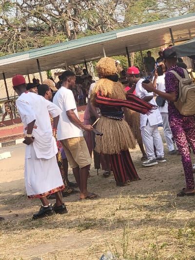 The image captures an outdoor cultural event in San Martín, Colombia, under bright daylight. A central figure is featured in an elaborate costume composed primarily of light brown, straw-like material, forming a bushy headpiece and a skirt over dark, red-and-black vertically striped trousers. Two strips of red and black striped fabric are wrapped around the upper torso of this figure, who also holds a dark stick in the right hand.
To the immediate left, a man in traditional white attire, consisting of a tunic over trousers and a red cap, stands with his hands clasped behind his back, wearing black shoes. Next to him, another man wears a white t-shirt and tan shorts, appearing to wear flip-flops, and holds a dark stick. To the far right, a man dressed in a patterned purple long-sleeved shirt and matching trousers, wearing a black baseball cap and a light-colored backpack, is actively recording the scene horizontally with a smartphone.
Multiple other individuals are visible in the background, some in similar white traditional attire with red caps, others in various casual clothing, observing or recording the event. The ground consists of dry earth with sparse, light brown grass. In the mid-ground, a low, long building with a light-colored roof structure is visible, and a large tree with mostly bare branches dominates the distant background.