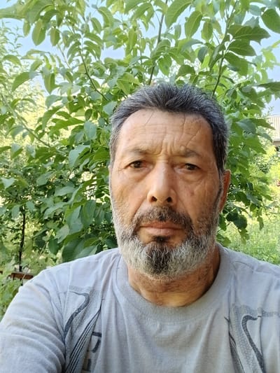 A medium shot shows a man with graying hair and a beard looking directly at the camera. He is wearing a light gray t-shirt with a dark graphic design on the chest. The man is outdoors, with lush green foliage and trees in the background. The lighting suggests it is daytime, with natural sunlight illuminating the scene. The image appears to be a selfie, taken from a slightly low angle. The location is described as Ghusar, Tajikistan.
