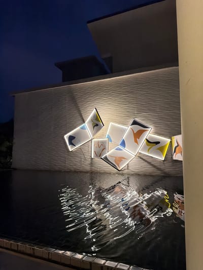 An exterior shot captures a modern building facade at night, featuring a textured wall with a series of illuminated rectangular art pieces. These pieces are mounted on the wall and display abstract colorful designs. Below the wall, a dark pool of water reflects the light from the art and the surrounding environment, creating shimmering patterns on its surface. The architectural elements suggest a contemporary design, with clean lines and a minimalist aesthetic. The lighting creates a dramatic ambiance, highlighting the textures of the wall and the artistic elements. The scene is devoid of any people or visible activity. The overall impression is one of serene, artistic nighttime illumination in an urban or resort setting, potentially in the city of Bang Muang, Thailand, given the context provided.