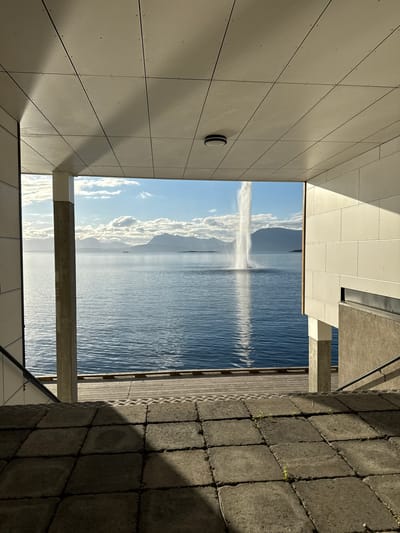 This image captures a scenic view of the harbor in Harstad, Norway, as seen from under a modern architectural overhang. The foreground is dominated by rough, gray paving stones with patches of green weeds growing between them, leading down a set of concrete stairs with a metal handrail. Beyond the stairs, a wooden boardwalk borders a tranquil body of blue water. In the distance, a tall water fountain sprays upwards, creating a dramatic vertical element against the horizon. The background features a range of majestic, silhouetted mountains under a bright blue sky dotted with fluffy white clouds. The overall scene is one of serene natural beauty juxtaposed with contemporary urban design, bathed in the warm light of what appears to be late afternoon or early morning.