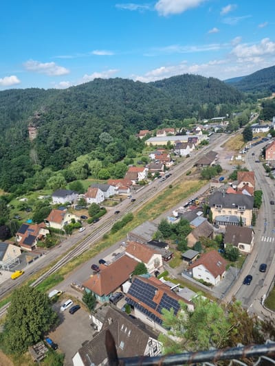 This is an aerial, long shot, daytime, eye-level, medium-wide, vertical, full-shot photograph of a residential area in Dahn, Germany. The scene is dominated by a dense, dark green forest that covers a steep hill on the left side of the image. A clear, bright blue sky with a few wispy white clouds is visible in the upper left quadrant.
To the right of the forest, a railway track runs diagonally across the frame. Adjacent to the railway, a road is visible with several cars parked along its edge and a few moving. A scattering of residential buildings, mostly with red-tiled roofs, are nestled between the forest, the railway, and the road. Some of these buildings have solar panels installed on their roofs. The houses appear to be of various sizes and architectural styles, with gardens and trees surrounding them. The overall setting suggests a semi-rural or suburban environment where nature and human habitation are in close proximity. There is no discernible activity or event taking place; the image captures a still moment. The weather appears to be fair and sunny, indicating a clear day. No text is visible in the image.