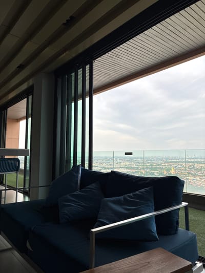 The image depicts a modern, open-air seating area on a high-rise balcony overlooking a sprawling cityscape, likely within the vicinity of Sao Thong Hin Town Municipality, Thailand. The foreground features a dark blue, low-profile sofa with several matching cushions, resting on a synthetic green turf floor. A metallic armrest extends across the sofa. To the left, a partial view of a white table and a blue woven chair is visible. The background is dominated by a vast urban landscape, stretching towards a hazy horizon under an overcast sky. Large, tinted glass panels form a barrier at the edge of the balcony, offering an unobstructed view of the city, with buildings of varying heights and architectural styles spread across the terrain. The architecture of the seating area itself suggests a contemporary design, with a slatted wooden ceiling overhead and dark-framed glass sliding doors that appear to be partially open. The overall atmosphere is one of a serene, elevated vantage point, providing a panoramic perspective of urban life.
