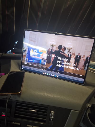 An overhead, slightly angled shot depicts the interior of a vehicle, likely a car, in the city of Dessau, Germany. A silver-edged tablet or portable monitor displays video content horizontally, resting on the dark gray, textured dashboard. The video shows a split screen: on the left, an advertisement for "ЕКЗО-ДЕРИЛ МАКСИ ЛАК" (EXO-DERIL MAXI LAC), a nail treatment, with a blue box and bottle visible, along with Bulgarian text stating "ЛЕCНО РЕШЕНИЕ ЗА ЗДРАВИ НОКТИ" (EASY SOLUTION FOR HEALTHY NAILS) and instructions for weekly application. On the right, a man and woman are depicted ballroom dancing in a formal setting, possibly a grand hall, with other formally dressed individuals and chandeliers in the background. Below the video, a progress bar, playback controls, and a "SANDOZ" logo are visible, along with fine print in Bulgarian concerning the product. To the left foreground, a dark-colored smartphone, possibly with a copper-toned case, lies flat on the dashboard, connected by a black charging cable. Below the smartphone, car air vents with horizontal slats and a chrome trim are present, with a red illuminated "P" indicator light and other car controls partially visible further down. The upper portion of the image shows dark fabric with thin red and light gray vertical stripes, likely part of the car's interior trim or headliner.