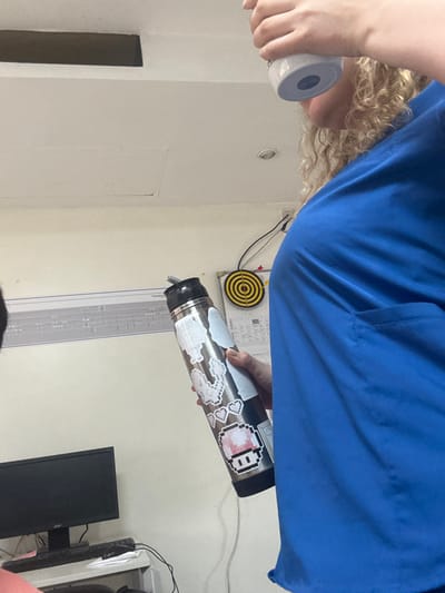 An indoor, close-up shot, originally rotated clockwise, captures a person from the chest up, engaged in a pouring action. The individual has curly blonde hair and is wearing a blue shirt, possibly scrubs. Their right hand holds a metallic water bottle, heavily decorated with various pixel art stickers, including a distinct white character with pink cheeks and several cloud-like designs. Liquid is actively being poured from this bottle into a transparent container with visible measurement markings, positioned below. The person's left hand holds a white, cylindrical object, possibly a roll of tape or a small container.

The background reveals a light-colored wall. A small yellow and black dartboard is mounted on the wall, with wires running nearby. A recessed light fixture is visible on the upper wall/ceiling, and a small rectangular door or panel is present on the wall to the left. A black monitor screen is partially visible on the right side of the frame, suggesting a workspace setting. The location context for this image is Adrogué, Argentina. No explicit text is clearly legible within the frame, beyond the pixel art designs on the water bottle.