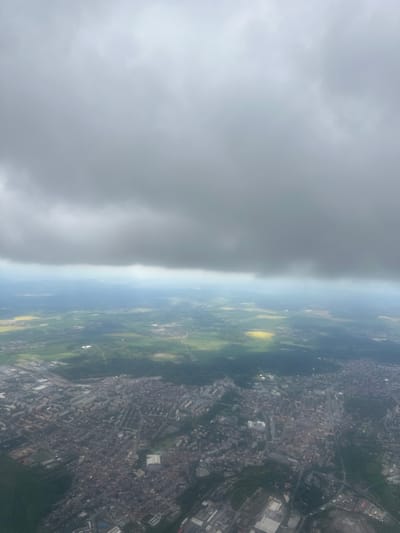 The image depicts a view from an airplane approaching Kladno, Czechia. The right side of the image shows the city's urban layout, including buildings and roads. To the left is a distinct cloud formation. Fields of green and yellow cut through the middle.