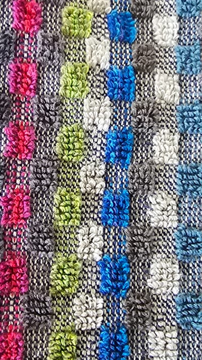 The image shows a close-up view of a textured fabric with a repeating pattern of raised, tufted loops. The loops are arranged in vertical columns and are colored in alternating stripes of bright pink, gray, lime green, white, royal blue, and light blue. The background fabric appears to be a dark navy blue with a subtle white flecked weave. The lighting suggests it is an indoor setting, with the texture of the fabric highlighted by soft illumination. There are no people, animals, or any recognizable landmarks visible in the image that would indicate a specific location in Vöhringen, Germany. The scene is purely a detail shot of textile material.