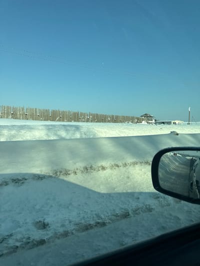 The image depicts a winter scene from a vehicle's perspective. A snow-covered road runs alongside a fence, with occasional buildings in the distance under a clear blue sky. The vehicle's side mirror is visible, reflecting the road and a hint of another car. Snow covers the ground and the road surface, with dark tracks indicating tire marks. The scene suggests a cold, possibly rural, environment.
