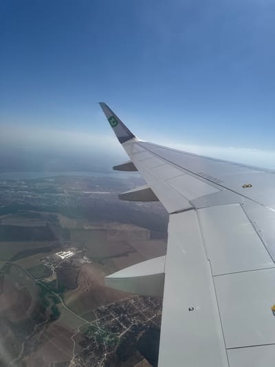 An aerial view from an aircraft window shows the right wing of the plane extending from the mid-right towards the top. The wing is white, with visible control surfaces and a winglet featuring a green 't' logo within a circle, indicative of TUI fly. The number "2" is marked on a section of the wing. Below and to the left of the wing, the landscape below consists of cultivated fields, some barren brown and others with green vegetation, intersected by dirt roads. A small settlement with dispersed buildings and roads is visible in the immediate bottom-left. Further in the distance, a larger expanse of developed land with denser housing, likely the city of Vaglen, Bulgaria, or its immediate surroundings, is present, alongside sections of forest or woodland. A significant body of water, appearing as a lake or large reservoir, stretches across the distant horizon, partially obscured by atmospheric haze. The upper portion of the image displays a clear, bright blue sky. The scene is illuminated by daylight.