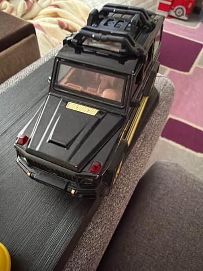 A black toy car, resembling a G-Wagon, is positioned on a grey, textured surface. The car has gold accents and red taillights. In the background, a red toy vehicle is partially visible, along with a pink and purple rug and a beige blanket. The scene is indoors, with no indication of the city of Mirnoye, Ukraine.