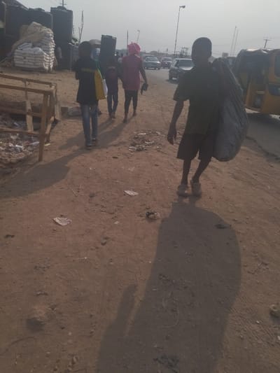 The image shows a street scene in Way, Nigeria, located at latitude 9.029 and longitude 7.584. Four people, including two adults and two children, are walking on a dirt road. The adult female, wearing a pink shirt and a pink hat, is walking in front of the other three. Two of the children are wearing black shirts, and the other child is wearing a green shirt. The child in the green shirt is carrying a large black bag over their shoulder. There are multiple vehicles parked on the side of the road, and a bus can be seen in the background. The road is lined with debris, and there are large blue water tanks and other debris in the foreground. The scene is bathed in warm sunlight, and the shadows of the people are long and thin.