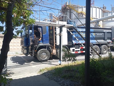 A blue and black Migmotor dump truck is parked on a paved road in the city of Ararat, Armenia. A man wearing black clothing is standing on the driver's side steps of the truck, partially visible through the open door. The truck is adjacent to a large industrial facility with elevated walkways and structures. The sky is clear and blue. A tree with green leaves and seed pods is in the foreground on the left. The ground is covered in asphalt and gravel.