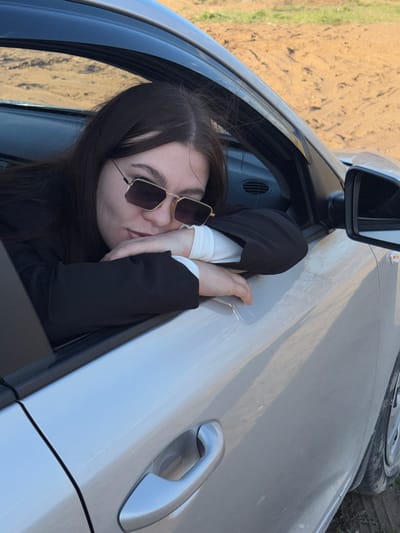 A young woman with long, dark hair and sunglasses is leaning out of the open window of a silver car. She is wearing a white shirt with a black jacket. The car appears to be parked on the side of a road, with a dirt field visible in the background. The location is likely Zaosinovo, Russia, as requested.