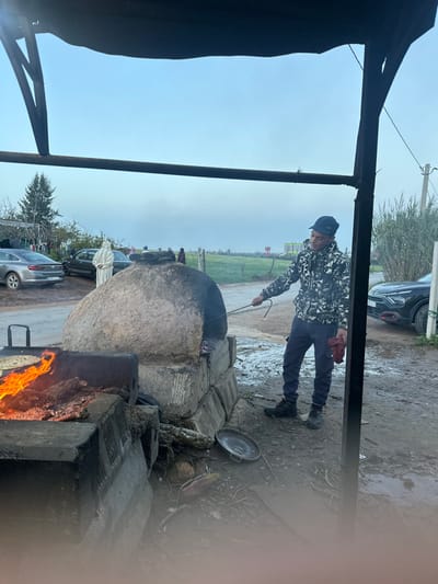 A man in a patterned jacket and blue pants tends to an outdoor oven. He holds a long metal tool, possibly adjusting the fire. The oven appears to be made of concrete or stone, with fire blazing in front. A black pan rests nearby. The scene is outdoors, with a cloudy sky and greenery visible in the background. Parked cars are also visible. The image seems to be taken from inside a structure, with a partial frame visible on the left.