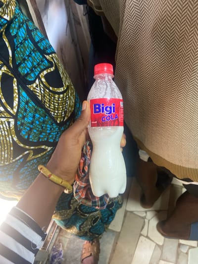 A woman is holding a bottle of Bigi Cola in Okengwe - Obehira, Nigeria. The bottle is red, white, and blue. The woman is wearing a patterned blue and black fabric over her lap. She has a yellow bracelet on her left wrist. In the background, there are other people and tiled floors. The floor is light gray. The surface under the bottle of Bigi Cola is covered in a red, blue, white and tan patterned fabric. The shirt that the woman has on is black and white striped. The Bigi Cola bottle is covered in condensation.