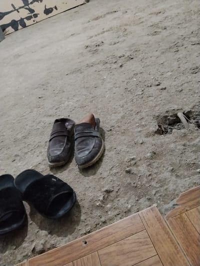 The image depicts a pair of dark brown leather loafers and two black slippers on a rough, gray concrete floor. The loafers are positioned side-by-side, with one appearing slightly more worn than the other. The slippers are located in the foreground, to the left of the loafers. The concrete floor has some debris and a small hole in the upper right section. A wooden tiled floor is visible in the bottom right corner. The background features a wall with dark, splattered paint. The scene appears to be an interior space in a dwelling. There is no indication of the city of Dagestanskiye Ogni, Russia, in the image itself.