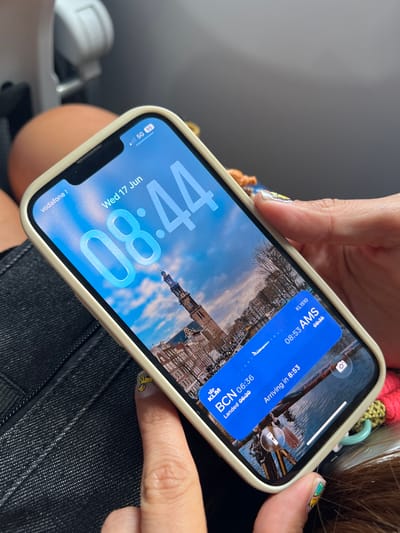 An iPhone 14 Pro displaying a flight information widget is held by a person's hands. The widget indicates a KLM flight from Barcelona (BCN) which landed at 06:36 and is arriving in Amsterdam (AMS) at 08:53. The phone's background is a cityscape with canals and buildings, likely Amsterdam, Netherlands. The time on the phone screen is 08:44. The phone is in a light-colored protective case. The person's fingernails have colorful, cartoonish designs. The phone rests on dark-colored denim fabric, possibly jeans. The image appears to have been taken from a slightly elevated perspective, suggesting the person is seated. The overall environment appears to be indoors or in transit, possibly on an airplane, given the background elements like the headrest. The city of Hoofddorp is located near Amsterdam and Schiphol Airport, a major hub for KLM flights.