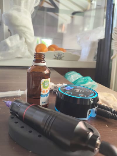 The image displays a collection of items commonly used for tattooing, arranged on a wooden surface. In the foreground, a black tattoo pen with a purple tip rests on a black stand. To the right, a compact, circular power supply unit with a blue illuminated rim and a digital display is visible. The unit is labeled "AURORA II" and features an omega symbol. Two cables are plugged into the back of the power supply. Behind the power supply, two teal-colored packets, possibly containing sterile wipes or disposable covers, are present. To the left of the power supply and in front of a brown glass bottle, a white syringe lies on its side. The bottle, which appears to contain a clear liquid, has a white cap and a colorful label with a floral design and the text "40 мл". In the background, an out-of-focus interior scene is visible, suggesting an indoor setting. A white, draped object is prominent, and behind it, a shelf holds several oranges. The overall lighting suggests it is daytime. The context provided indicates the location is Melikhovo, Russia.