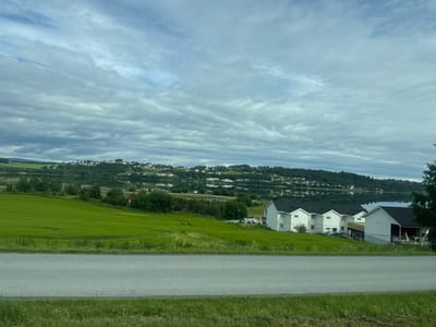 The image shows a rural landscape in Levanger, Norway, with a body of water in the background, a green field in the middle ground, and a road in the foreground. The sky is overcast with gray and white clouds. On the far side of the water, a town or village is visible on a hillside, with many houses. In the middle ground, there are several modern white houses with dark roofs situated on the edge of the green field. The field is lush and green, with some unevenness in its surface. The road is paved and runs horizontally across the bottom of the image. Grass and some vegetation are visible along the edges of the road. The overall scene depicts a serene and somewhat residential area surrounded by natural scenery.