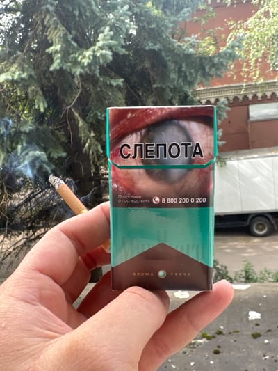 A person's hand holds a lit cigarette, emitting smoke, and a pack of cigarettes. The cigarette pack is turquoise and brown and features a graphic of an eye with a clouded pupil, and the word "СЛЕПОТА" (blindness) in large black letters. Below this text, smaller text reads "Подробнее о последствиях" (More about the consequences) and a phone number, "8 800 200 0 200." At the bottom of the pack, the words "AROMA FRESH" are visible. The background consists of a dense evergreen tree and a blurred building and white truck. The overall scene suggests an outdoor setting, possibly a city environment given the presence of the truck and building. The image was taken in Dedovsk, Russia.