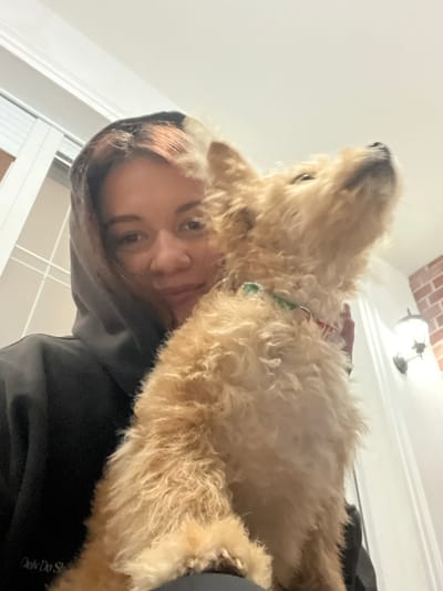 A low-angle, slightly tilted, selfie captures a person in a black hooded sweatshirt alongside a golden doodle dog. The person's face is visible under the hood, with light brown hair peeking out. The dog, mostly in focus, has fluffy, light golden fur. A collar is visible around the dog's neck. In the blurry background, a white-painted wall with a door and a light fixture on the ceiling can be seen.