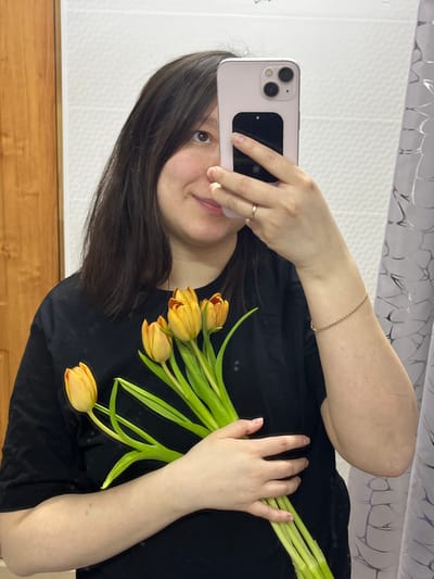 An indoor mirror selfie taken in a residence located in Kozlovka, Russia, depicts a female subject from the waist up. The subject, with dark brown, shoulder-length hair and a light complexion, wears a plain black short-sleeved t-shirt. She is captured with a slight smile, her left eye visible while her right eye is partially obscured by a light pink-cased smartphone, which she holds in her left hand to take the photograph. A simple, light-colored ring is present on the fourth finger of her left hand. Her right wrist displays a thin, light-colored chain bracelet. In her right hand, held against her body, is a bouquet consisting of approximately five to seven yellow-orange tulips with prominent green stems. The background consists of a textured white tiled wall on the left, a vertical light brown wooden panel or door frame, and a white or light-colored curtain with a subtle patterned design on the far right.
