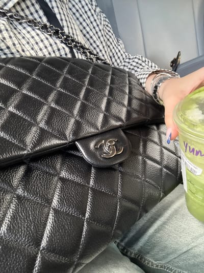 Here is a detailed description of the image:
The image presents a close-up view of a person holding a green-colored beverage, possibly a smoothie, over a black, quilted leather bag. The bag is secured with a metal clasp featuring a recognizable logo. The person is wearing multiple silver bracelets and a black and white gingham patterned shirt. The background is soft and slightly blurred, revealing light grey denim and a light grey surface. The focus is sharp on the bag and the hand holding the drink. The scene conveys a casual, everyday moment. The location, Chula Vista, United States of America, cannot be determined from the image alone.