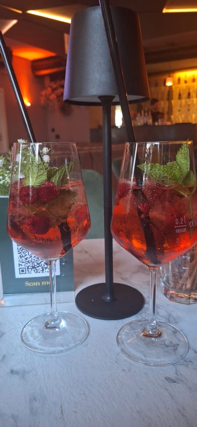Two glasses of Aperol Spritz, garnished with mint and raspberries, are displayed on a marble table. The glasses are tall and clear, with a wide base. A black lamp with a conical shade stands behind the glasses, and a blurred bar scene with bottles and lights is visible in the background. The overall ambiance suggests a bar or restaurant setting. The image does not contain explicit information about its location in Neuwied, Germany.