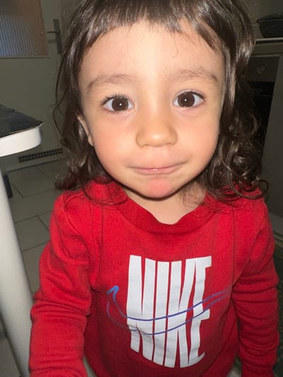 A young child with dark, curly hair and brown eyes is looking directly at the camera, making a slight pout with their lips. The child is wearing a red long-sleeved shirt with a white Nike logo on the front. The background is out of focus but appears to be an indoor setting, possibly a kitchen or hallway, with a door and a counter visible. The image was taken in Fulda, Germany.