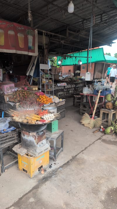 The image depicts an outdoor food stall in Phao, Laos, featuring a variety of food items and cooking equipment. In the foreground, a large black pot sits on a makeshift stove constructed from a yellow crate and concrete blocks, with skewers of food being cooked and steaming. Various fruits, including rambutan and oranges, are displayed in crates behind the cooking area. In the background, two young men walk away from the stall, and a green tarp provides shade over part of the scene. The environment appears to be a street or market area, with a concrete ground and a rustic wooden roof structure overhead.