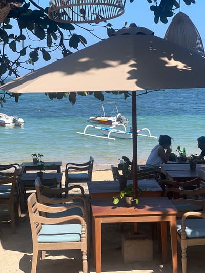 An outdoor dining area is set up on a sandy beach overlooking a turquoise ocean. Several wooden tables and chairs with light blue cushions are arranged under a large, light-colored umbrella. The tables are set with potted plants. In the background, two boats are anchored in the water, one a small white motorboat and the other a traditional outrigger canoe. Two individuals are seated at a table in the mid-ground, facing away from the camera. One person is wearing a white tank top and the other is wearing a dark shirt. The sky is clear and blue, and the sun casts shadows from the umbrella and overhead foliage onto the scene. The location is identified as Denpasar, Indonesia.