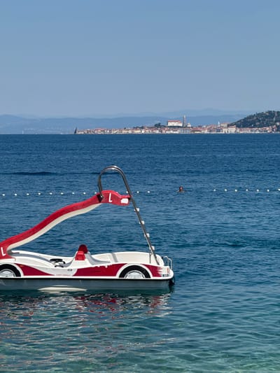 A pedal boat, designed to resemble a red and white sports car with faux wheels, is moored in clear, blue ocean water. The boat has a red slide extending from its front. A person is visible swimming in the water behind the pedal boat, closer to a white buoy line. In the distance, a coastal city with red-roofed buildings and a prominent church tower is situated on a hill. The sky is clear and blue. The location is the coast of Crveni Vrh, Croatia.