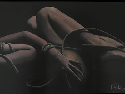 The image is a close-up, low-angle shot of a person's body, bound with thick rope. The person is lying down, with their legs bent and their arms crossed across their torso. The rope is tightly wrapped around their wrists and forearms, and also appears to be securing their legs. The lighting is dim, casting long shadows and highlighting the contours of the body. The skin tone is a warm brown, and the rope is a natural fiber, likely jute or hemp. There is a signature in the lower right corner of the image, which appears to read "P. Panteloye." The background is a dark, nondescript surface. There is no specific indication of location in Mesogi, Cyprus.