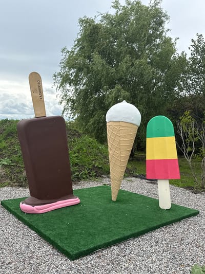 This is an outdoor, eye-level, medium shot of three oversized ice cream sculptures placed on a rectangular green artificial turf mat, which sits on a bed of grey gravel. The backdrop consists of a grassy hill with scattered foliage and trees under a cloudy sky. To the left, a large, brown chocolate bar ice cream sculpture with a pink bottom is positioned diagonally, leaning slightly to the right. The wooden stick of this sculpture is visible at the top and features the word "КАЛИНКОВО" in Cyrillic, along with a floral emblem. In the center, a tall ice cream cone sculpture with a white scoop of ice cream stands upright. To the right, a brightly colored popsicle sculpture, divided into green, yellow, and red sections, is positioned upright on its white stick. No people are present in the image, and there are no discernible actions or interactions occurring. The overall scene depicts a public art installation or decorative display, likely in Kalinkovo, Russia, given the inscription on the sculpture.