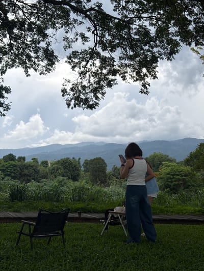 The image depicts an outdoor scene in Tambon San Phi Suea, Thailand, under a cloudy sky. In the foreground, a black camping chair sits on a grassy lawn. A wooden walkway runs behind the chair, leading to a low table where some items are placed, including what appears to be a beverage in a cup. Two individuals are standing on the walkway. One person, viewed from the back, wears a white tank top with red trim and dark blue, wide-leg pants, and is looking at a mobile phone. Another person is partially visible to their right, wearing a light blue garment and white shorts. The background features lush green vegetation, including tall grasses and trees, with a range of mountains visible in the distance. Overhead, dark branches and leaves from a large tree frame the upper part of the image.