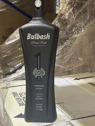 The media file is an image showing a dark-colored bottle of Bulbash Black Pearl premium vodka. The bottle has a distinctive shape, wider at the base and tapering towards the neck. Text on the bottle indicates "Bulbash Black Pearl Finest Quality," "Premium Vodka," "Limited," "Product of Belarus," and "Black Pearl Filter." A circular emblem with the number "1" and the brand name is also visible. The bottle appears to be wrapped in clear plastic, suggesting it might be in storage or awaiting shipment.
In the background, there are cardboard boxes, some of which have markings. One box has the text "HANDLE WITH CARE" printed on it. Another box has an arrow and the word "HANDLE" visible. The lighting in the scene suggests an indoor setting, possibly a warehouse or storage area, with ambient light. There are no people visible in the image. The overall scene is focused on the product and its packaging, with hints of a commercial or logistical context. The location context provided is Zaslaŭje, Belarus, which aligns with the "Product of Belarus" text on the bottle.