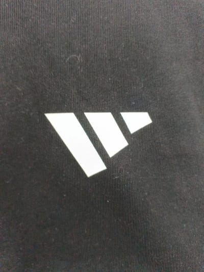 This media file is a close-up shot of a black fabric, likely clothing, with a white logo. The logo consists of three slanted parallelograms arranged to form a triangular shape. The fabric has a visible ribbed texture. There is no text present in the image. The lighting is neutral, and no specific time of day or weather conditions can be inferred. The setting is a simple, unidentifiable surface, emphasizing the logo itself. No people or specific location cues are visible beyond the product's origin in Znamya Oktyabrya, Russia.