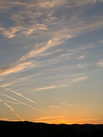 This is a vertical, wide shot of a sky at sunset over Svinjišta, Montenegro. The sky is a gradient of colors from a pale orange near the horizon to a light blue at the top. There are thin, wispy clouds scattered throughout the sky, tinged with orange from the setting sun. Several white contrails from airplanes are visible, streaking across the sky in different directions. The bottom of the image features a dark, silhouetted landscape, likely hills and trees, against the vibrant sunset. There are no people or animals visible in the image.