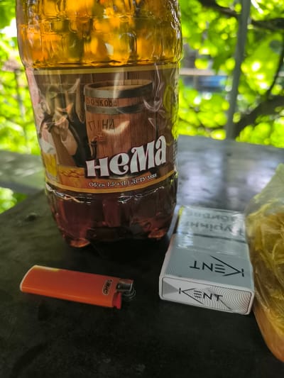 A close-up shot, taken outdoors, features a plastic bottle filled with a golden-amber liquid, likely beer, positioned slightly left of center. The bottle's label depicts a wooden barrel with the text "БОЧКОВЕ" and "ПІНА" in Cyrillic script, along with the brand name "Нема". Below this, text reads "Об'єм 1,5 л (1) ДСТУ 3888". To the right of the bottle, a pack of Kent cigarettes is visible, its silver packaging featuring the brand logo. An orange BIC lighter lies to the left of the cigarette pack. The objects rest on a dark, textured surface, possibly a table. In the background, out-of-focus green foliage suggests a natural, outdoor setting. The image captures a still life arrangement of common consumables, with the environment indicating a location in Bohodukhiv, Ukraine, possibly in a park or backyard.
