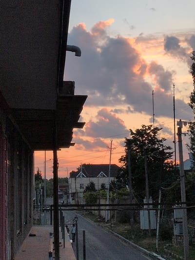 The image depicts a dusky sky with dramatic clouds illuminated by the setting sun. The sky is filled with hues of orange, pink, and grey, creating a vibrant contrast with the darker elements in the foreground and midground. In the lower foreground, the silhouettes of rooftops and a chimney are visible, suggesting an urban or semi-urban environment. A white pipe protrudes from one of the rooftops.

Further back, the scene transitions to a line of trees and utility poles with numerous wires stretching across the frame. The silhouette of a building with a triangular roof is discernible amongst the trees, hinting at residential or commercial structures. The road is visible on the right side, appearing to be asphalted. The overall atmosphere is serene and somewhat melancholic, characteristic of a sunset in a populated area. The time of day is clearly evening, as indicated by the low sun and the colors in the sky. There is no visible text in the image. The setting appears to be Şəki, Azerbaijan, suggested by the architectural style of the building with the triangular roof.